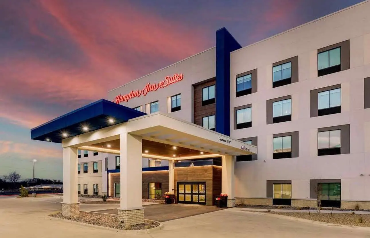 A Hampton Inn & Suites at sunset, its blue-roofed porte-cochère lit beneath the red script sign